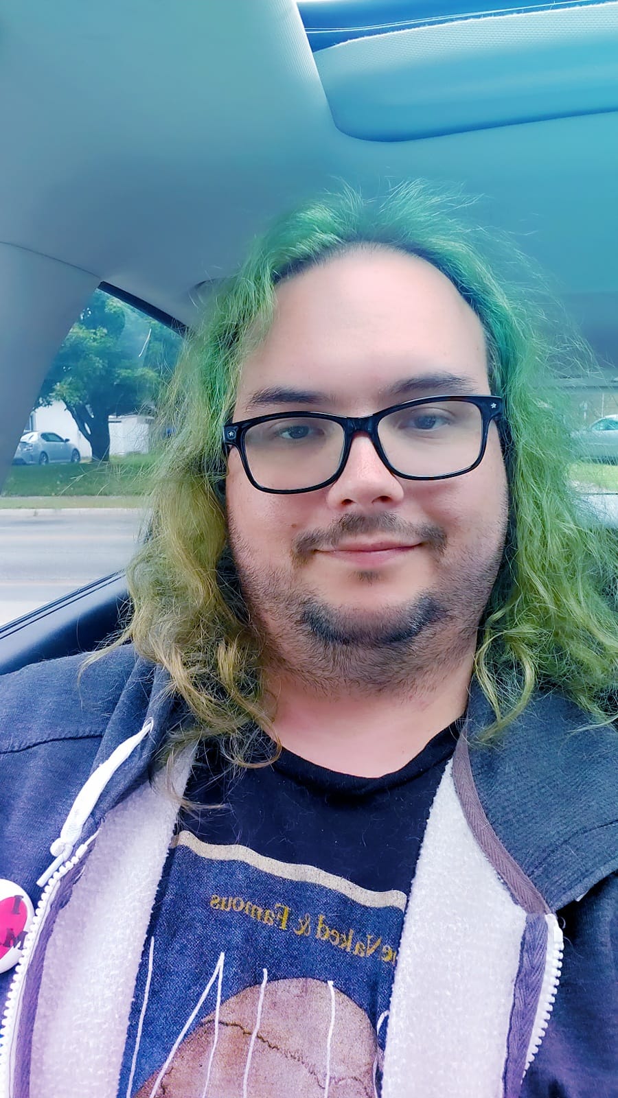 A man with long green hair wearing a hooded sweater vest sits in a car